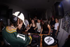 DJ im grünen Trikot im Vordergrund, davor tanzende Menge im Club.