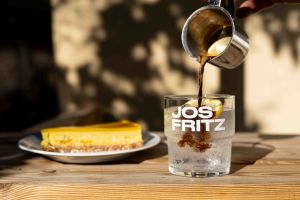 Eine Hand gießt Espresso aus einem kleinen Metallkännchen in ein Glas mit Eis und Zitrone, das die weiße Aufschrift "JOS FRITZ" trägt. Im Hintergrund steht ein Teller mit Kuchen.