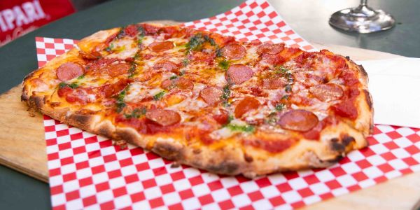 Eine fertige, rechteckige Pinsa mit Tomatensoße, Käse und Salami liegt auf rot-weiß kariertem Papier auf einem Holztisch.