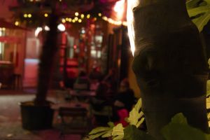 Baumstamm im Vordergrund, dahinter unscharfer Außenbereich mit Tischen bei Nacht.