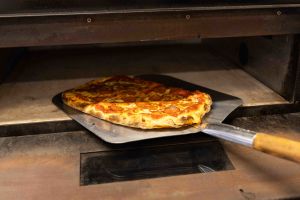 Eine fertig gebackene, rechteckige Salami-Pizza wird mit einem Pizzaschieber aus Holz und Metall aus einem heißen Ofen geholt.