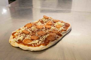 Eine rechteckige Pizza, belegt mit Tomatensoße, geschmolzenem Käse und Salamischeiben, liegt auf einer metallenen Arbeitsfläche.