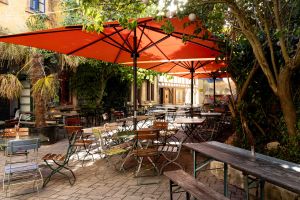 Ein sonniger Café-Innenhof mit mehreren Tischen und Stühlen aus Holz und Metall, überspannt von großen orangefarbenen Sonnenschirmen.