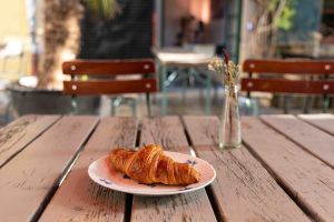 Ein frisch gebackenes Croissant liegt auf einem kleinen, weiß-blauen Teller auf einem Holztisch im Freien. Dahinter steht eine kleine Glasflasche mit Trockenblumen.