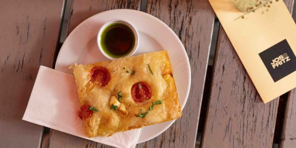 Ein Stück Focaccia mit Kirschtomaten und Kräutern auf einem Teller, serviert mit grünem Dip, einer Serviette und der „JOS FRITZ“-Speisekarte.