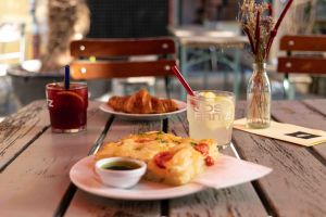 Ein reich gedeckter Holztisch im Außenbereich mit Focaccia und Dip, einem Croissant, zwei Erfrischungsgetränken im „JOS FRITZ“-Glas, einer Speisekarte und einer kleinen Blumenvase.