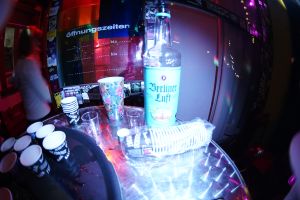 Flasche Berliner Luft und Becherstapel auf Tisch, bunte Beleuchtung im Hintergrund.