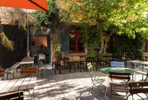 Der begrünte Außenbereich eines Cafés mit Holztischen, Bänken und Stühlen unter Bäumen vor einer Fassade mit rot gerahmten Fenstern.