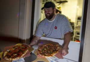 Lächelnder Mann reicht zwei Pinsas auf Holzbrettern durch ein Ausgabefenster.