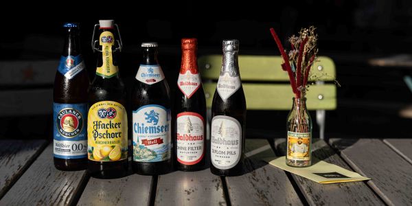 Sechs verschiedene Bierflaschen, darunter Paulaner, Hacker-Pschorr Radler, Chiemseer Hell und drei Sorten Waldhaus, stehen aufgereiht auf einem Holztisch im Freien neben einer kleinen Vase mit Trockenblumen und einer Speisekarte.
