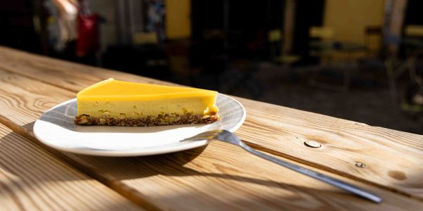 Ein Stück hellgelber Kuchen auf einem weißen Teller mit Gabel auf einem sonnenbeschienenen Holztisch im Außenbereich.