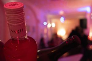 Flaschenhals einer Sarti-Flasche vor unscharfem, pink beleuchtetem Partyhintergrund.