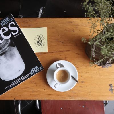 Eine Tasse Espresso auf einer Untertasse mit einem Löffel steht auf einem Holztisch. Daneben liegt ein Magazin mit „JOS FRITZ“-Stickern, eine gelbe Karte mit dem kreisförmigen Logo „L'ÉTÉ AVEC MARIE“ und eine Vase mit Trockenblumen.