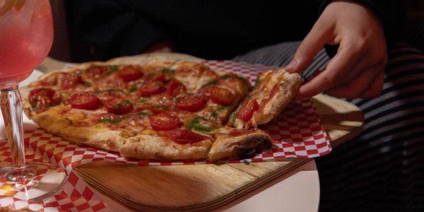 Hand nimmt ein Stück Pinsa vom Holzbrett, daneben ein pinker Drink.