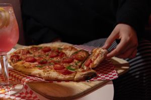 Hand nimmt ein Stück Pinsa vom Holzbrett, daneben ein pinker Drink.