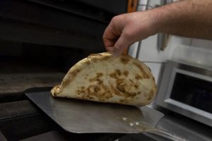 Hand schiebt eine Pinsa mit einem Pizzaschieber in den Ofen.