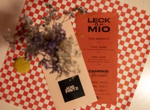 Speisekarte „Leck Oh Mio“, Trockenblumen und Jos Fritz Karte auf kariertem Untergrund.