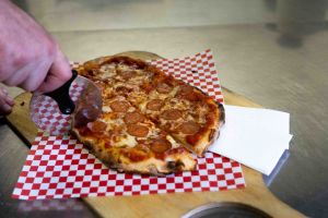 Eine Hand schneidet mit einem runden Pizzaschneider eine auf einem rot-weiß karierten Papier liegende Salami-Pizza in Stücke.