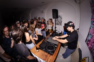 DJ rechts am Mischpult im Profil, umringt von Zuschauern.