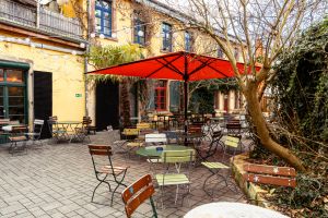 Außenbestuhlung unter rotem Schirm vor gelber Hausfassade.