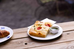 Ein Stück Focaccia mit Kirschtomaten und ein Schälchen mit grünem Dip stehen auf einem weißen Teller auf einem Holztisch, daneben ein Teller mit einem Croissant.