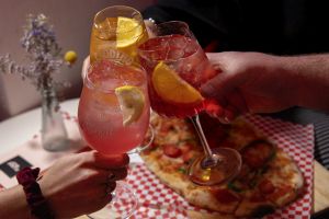 Drei Personen stoßen mit Spritz-Getränken über einer Pizza an.