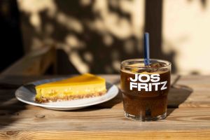 Ein Glas mit dem Aufdruck "JOS FRITZ", gefüllt mit einem Eiskaffee-Getränk, Eiswürfeln, Zitrone und Strohhalm, auf einem Holztisch neben einem Stück Kuchen.