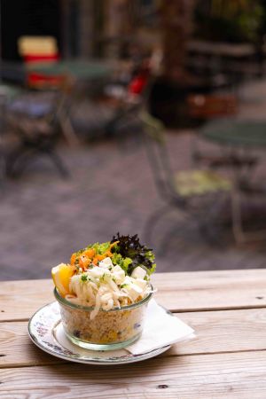 Eine kleine Glasschale, gefüllt mit einem bunten Salat aus Couscous oder Quinoa, Weißkraut, Karotten, Blattsalat und Fetakäse, garniert mit einer Orangenscheibe, steht auf einem Teller auf einem Holztisch im Außenbereich.